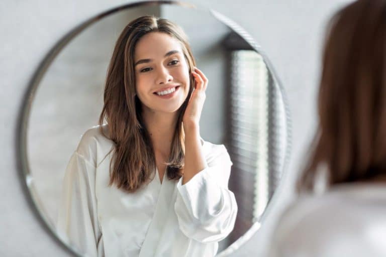 Woman with brunette hair and eyes looking into a bathroom mirror and smiling at herself. One hand is up against her cheek, lightly touching her long hair. The other hand is at her side and out of frame.  