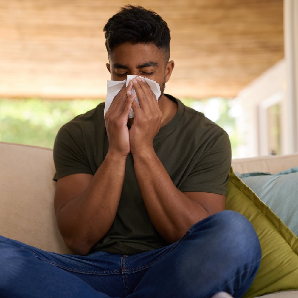 a man sits indoors on a couch and blows his nose